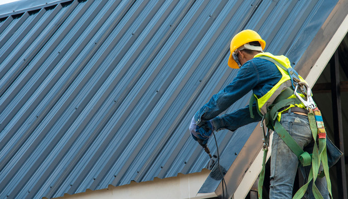 Construction worker wearing yellow helmet and safety harness belt during working on roof installation of metal roof of a house using air or pneumatic nail gun