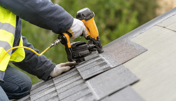 Roofer worker installing asphalt or bitumen tile on top of the roof under construction house using air or pneumatic nail gun