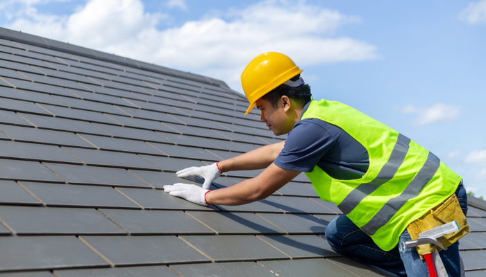 Roof repair worker with white gloves and a helmet replacing gray tiles or shingles on house with blue sky as background