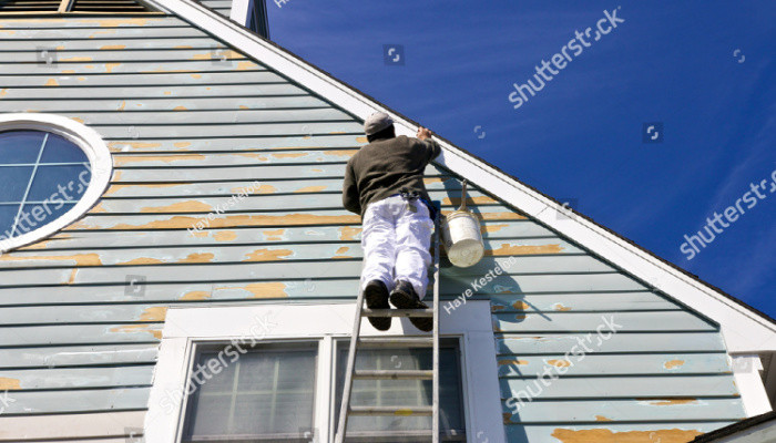 A contractor or painter in black shirt on a ladder doing exterior paint work on a house with wood siding with blue sky background