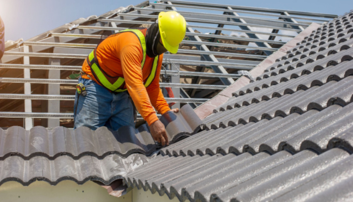 Roof repair worker with orange shirt and yellow helmet replacing gray tiles or shingles on house with blue sky as background