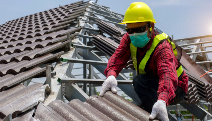 Roof repair, worker with white gloves and red shirt replacing gray tiles or shingles on house with blue sky as background
