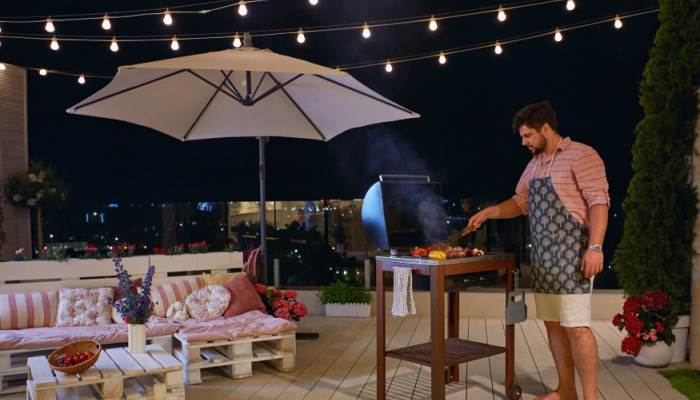 a man grilling barbecue on deck with string or rope lighting, enjoys warm summer night at home courtyard
