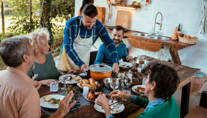 Man standing over the dining table with friends and family wearing a white apron, serving a pot with delicious food Group of diverse people for Holiday festive dinner party on the outdoor kitchen