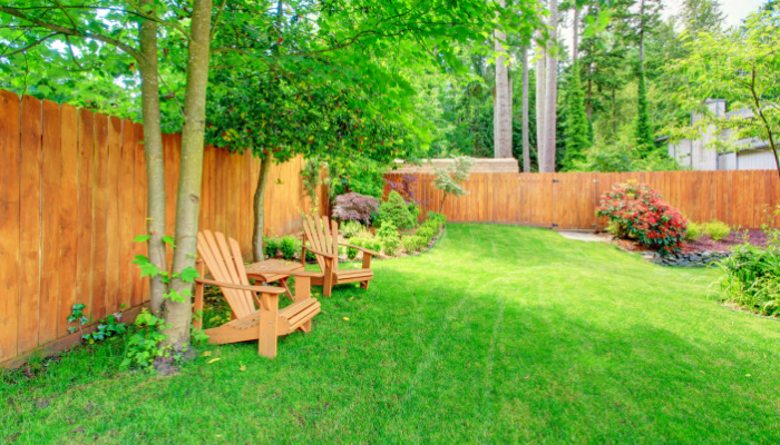 Fenced backyard with green lawn, flower beds and romantic sitting area with wooden chairs and table on a bright sunny day