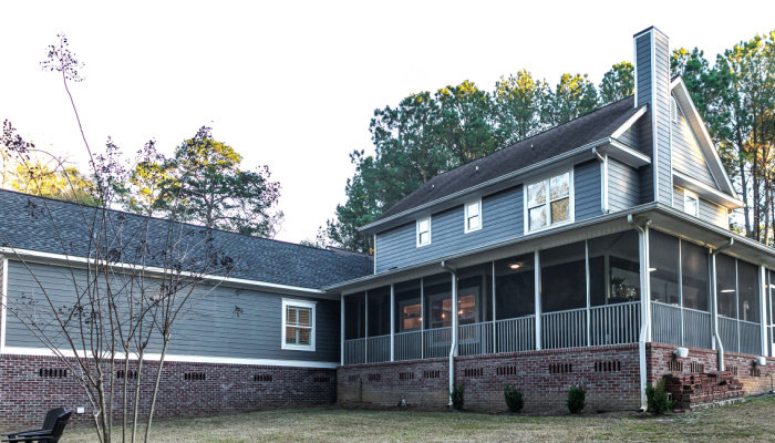 View of a large two story blue gray house with wood and vinyl siding and a large screened in porch patio with trees in the background