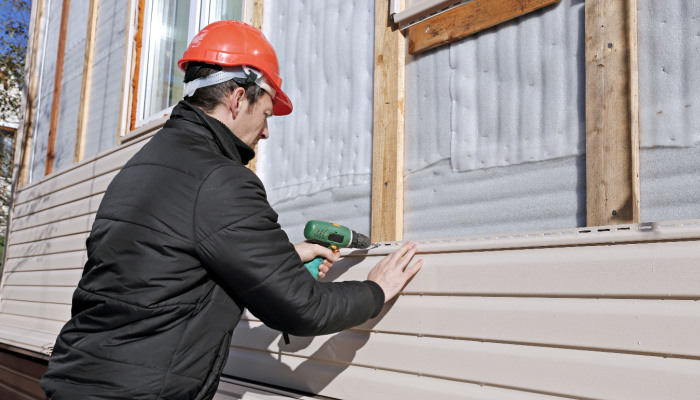 A worker with protective gear installs beige siding on the facade of the house